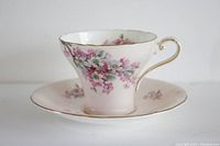 Teacup and saucer set with pink floral pattern and gold edging. The teacup shows front and side views highlighting the handle and floral design.
