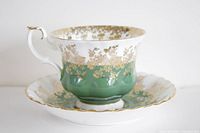 Side view of vintage porcelain teacup and saucer featuring green and gold floral pattern, scalloped edges, and gold trim.