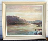 Full frontal image of framed landscape painting against white siding, showing river, mountains, trees, and sky with artist signature visible bottom left.