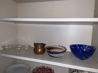 Shelf with multiple items: four clear glass bowl-like items, a copper container with two handles, a woven basket, blue and white swirl glass bowl, and a plate partially visible below.