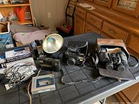 Overview of lot including vintage cameras, binoculars in leather case, cassette tape recorder and accessories arranged on black table.