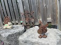 Photo showing two cat ornaments and two decorative wrought iron pieces placed on rock surface outdoors against a wooden fence background.