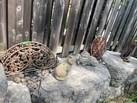 Photo showing the two cat statuettes placed on large stone surfaces in front of a wooden fence, with the semi-circular metal fence decorations behind them resting on the ground between stones.
