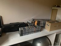 Two black metal CD racks filled with CDs next to a JVC stereo system, and a cardboard box full of CDs on a white shelf.