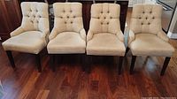 Four beige upholstered dining chairs with tufted backs, nail head trim, and dark wood legs arranged on wood flooring.