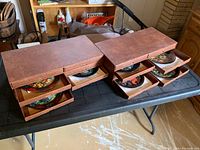 Two brown storage boxes with multiple drawers containing colorful collector plates, placed on black folding table.