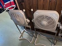 Pair of beige Air Innovations fans on stands with rectangular bases, showing front and side view in a basement area.