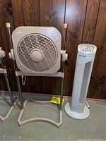 Air Innovations AI-4400 Airworks pedestal fan on stand and white tower fan side by side on floor with wood panel wall background.