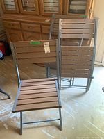 Three folding chairs arranged with two standing and one folded showing metal frames and faux wood slats.