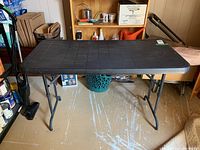 Front view of black plastic folding table on metal legs in a room with various items in the background.
