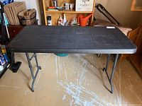 Full view of black plastic folding table with metal legs standing on a paint-spattered floor.