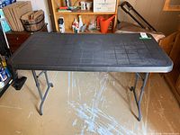 Full view of black plastic rectangular folding table with metal legs, top surface showing grid texture and wear marks.