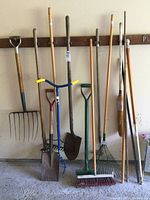 Photo showing full array of garden tools hanging on wall, including pitchfork, shovels, rakes, push broom and natural bristle broom.