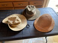 Three hats displayed on a table: leather fedora, straw pith helmet style, fabric wide-brimmed sun hat.