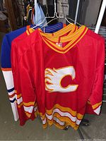 Three hockey jerseys hanging on wire hangers. Two red Calgary Flames jerseys in front with yellow and white striping and flaming 'C' logo. Behind is a blue Edmonton Oilers jersey with orange and white striping.