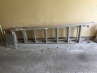 Photo of a 7-foot aluminum ladder lying horizontally on a concrete garage floor against a beige wall, showing full length and details of the rungs and base.