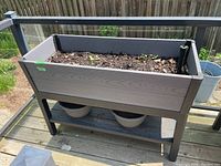 Full view of elevated rectangular resin planter with soil and small sprouting plants, standing outdoors on a wood deck with two plastic pots below.
