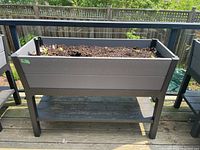 Front view of the self-watering raised planter showing soil with a few small plants and wood grain textured resin sides, standing on a deck.