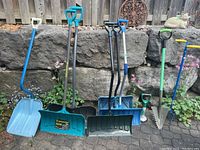 Wide view of six snow shovels, one garden shovel, and one garden claw lined up outdoors on stone patio against rock wall.