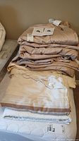 Stack of folded silk duvets in shades of beige and brown with labels visible, with folded beige blanket and white flannel sheet underneath