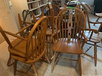 Six oak chairs shown grouped together in a room with bookshelves. Four without arms and two with armrests, natural oak wood finish, spindle backs with carved center splats.