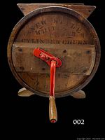 Front view of the wooden butter churn showing the circular churn body with the red metal hand crank handle and faded stenciled lettering.