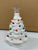Front view of white ceramic Christmas tree with colorful bulbs and clear star topper against plain brown cardboard background.