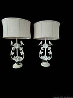 Photo of two cream-colored table lamps with bird and floral design bases and beige fabric shades against black background.