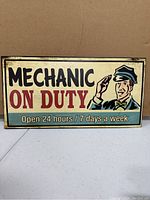 Photo shows a rectangular reproduction metal sign with a mechanic illustration and text 'MECHANIC ON DUTY Open 24 hours / 7 days a week'.