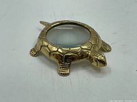 Brass turtle-shaped magnifier shown from a side angle highlighting turtle head and legs with clear glass lens at center.