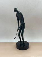 Full front view of bronze golfer sculpture showing elongated figure in mid-swing on round black stone base.