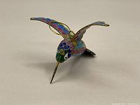 Small colorful cloisonné hummingbird figurine with wings outstretched photographed from front angle showing detailed enamel in blue, pink, purple, green, and gold.