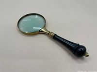 Full view of the vintage magnifying lens lying flat, showing brass rim and black handle with ornamental design.