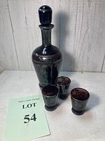 Full view of amethyst glass decanter with silver overlay and three matching shot glasses on a light wood surface with lot number card.