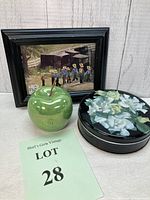 Framed Amish print, green glass apple and floral vintage candy tin arranged together on a white wood background.