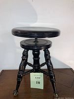 Front view of antique black wooden piano stool with round swivel seat and four turned legs ending in clear glass claw feet.