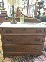 Full frontal view of the antique Eastlake marble top chest showing 3 wooden drawers with brass handles and wooden keyholes, sitting on a patterned rug in a room with various antique furniture.