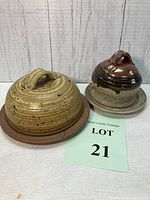 Front view of two ceramic butter/cheese dishes with covers on a white surface against a wood plank backdrop