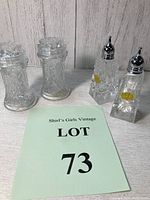 Photo showing both sets of vintage cut glass crystal salt and pepper shakers placed side by side on a wooden surface against a textured wall.