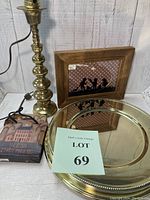 Photo showing brass lamp base with attached electrical cord, stacked gold metal chargers, framed shadow figure artwork, and 'Bless This Home' coasters and bag on table surface.