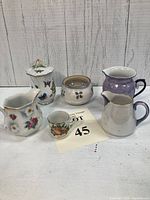 Overview of six small ceramic vases and creamers arranged on white surface with wood background, showcasing varying shapes, colors, and floral designs.