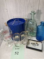 Complete lot showing all items including blue metal ice bucket with lid and wooden handles, three rocks glasses with gold trim, Miller beer glass with stem, two decorative bottles (clear and blue), and a box of hand-crafted wine glass charms.