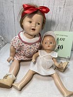 Front sitting view of the two dolls, smaller doll in white dress and bonnet, larger doll with red bow and cherry print dress