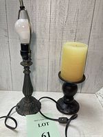 Two lamps side by side on a white surface in front of a wood panel wall. One lamp has an ornate black metal base with a visible light bulb and electrical cord with switch. The other is a black metal candle holder base with a thick yellow pillar candle on top.