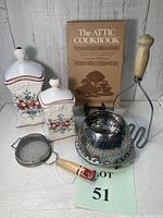 Photo showing the entire lot of vintage kitchen items including two Casafina canisters, The Attic Cookbook, metal colander, potato masher, and strainer.