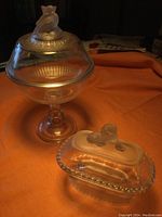Image showing both glass compotes on orange cloth background, large pedestal with bird lid and smaller rectangular with lion lid.