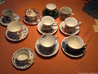 Full set of 10 bone china cups on saucers, various floral and scenic patterns shown on orange cloth background.