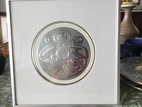 Frontal photo of embossed silver Haida World Drum artwork in a white frame with glass.