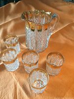 Overview shot showing all six items: one cut glass pitcher with gold wash trim and five matching cut glass drinking glasses with gold wash trims, arranged on an orange fabric background.