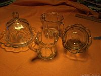 Photo showing butter dish with lid, spooner, creamer pitcher, and sugar bowl on orange cloth.